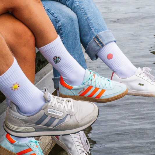 Weiße Tennissocken mit Sonnen-,Muschel-, und Monsterablatt-Motiv werden von 3 Personen präsentiert mit hellen Sneakern.