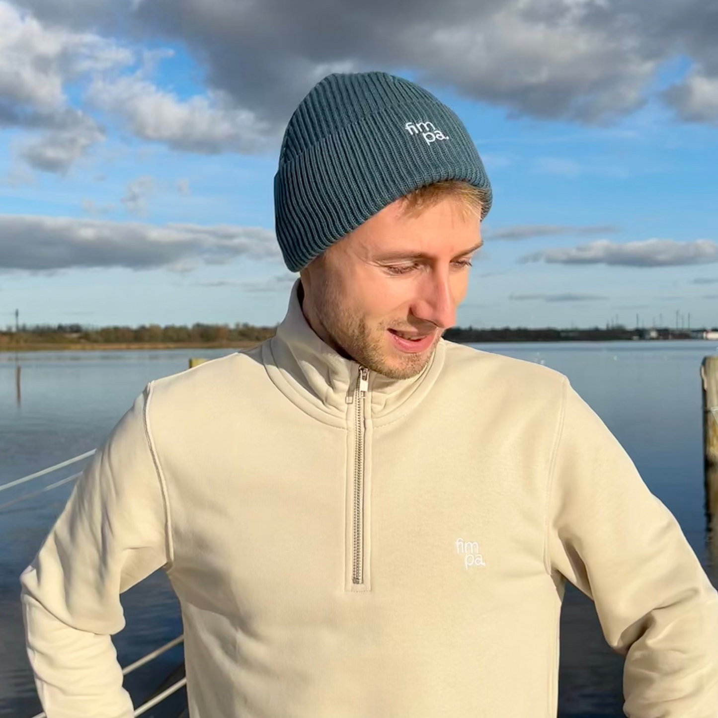 Person trägt einen petrolfarbenen fimpa-Beanie und einen beigen Pullover mit Reißverschluss vor einer malerischen Kulisse am Wasser – moderner und nachhaltiger Look.