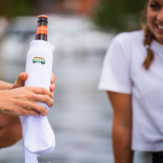 Eine weiße Bio Baumwollsocke umhüllt eine kühle Flasche Bier, im Fokus präsentiert, während eine Frau diese bewundert im Hintergrund.