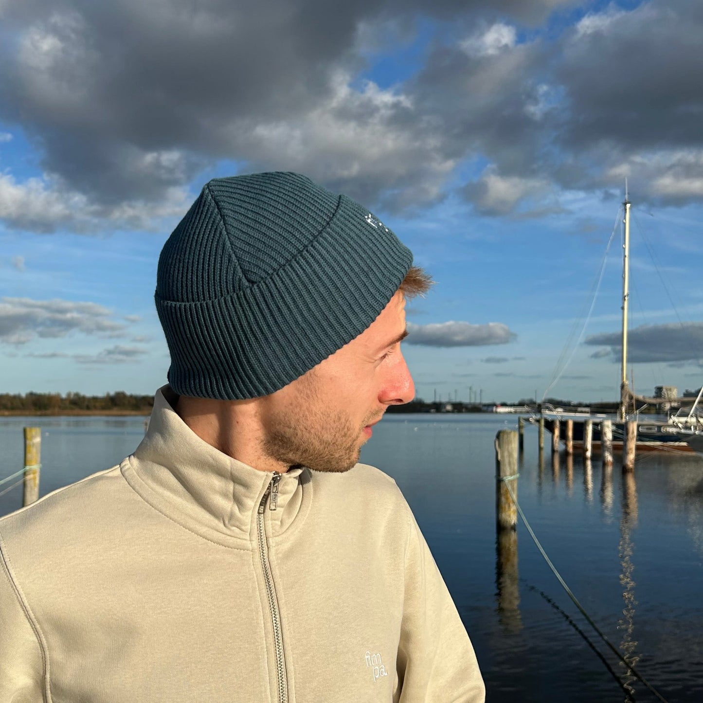 Person mit einem petrolfarbenen Beanie und einen beigen Zipper von fimpa vor einer ruhigen Hafenkulisse mit Segelbooten und blauem Himmel – lässiger und zeitloser Stil.