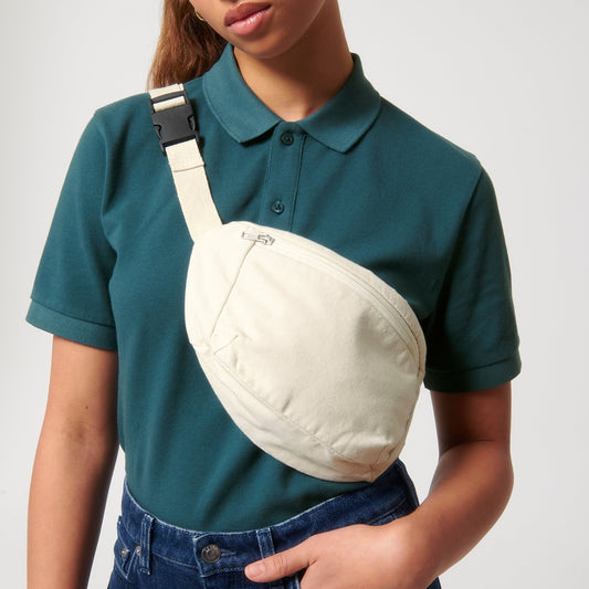 Frau in einem dunkelgrünen Poloshirt und blauer Jeans trägt eine cremeweiße Bauchtasche aus nachhaltigem Material quer über die Schulter. Die Tasche hat einen dezenten Reißverschluss und einen verstellbaren Trageriemen mit Clipverschluss.