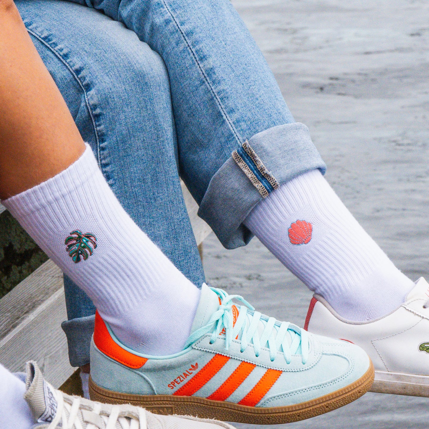 Weiße Socken mit gesticktem Monsterablatt-Motiv und Muschel-Motiv in grellen Farben und mit hellen Schuhen kombiniert am Wasser getragen.
