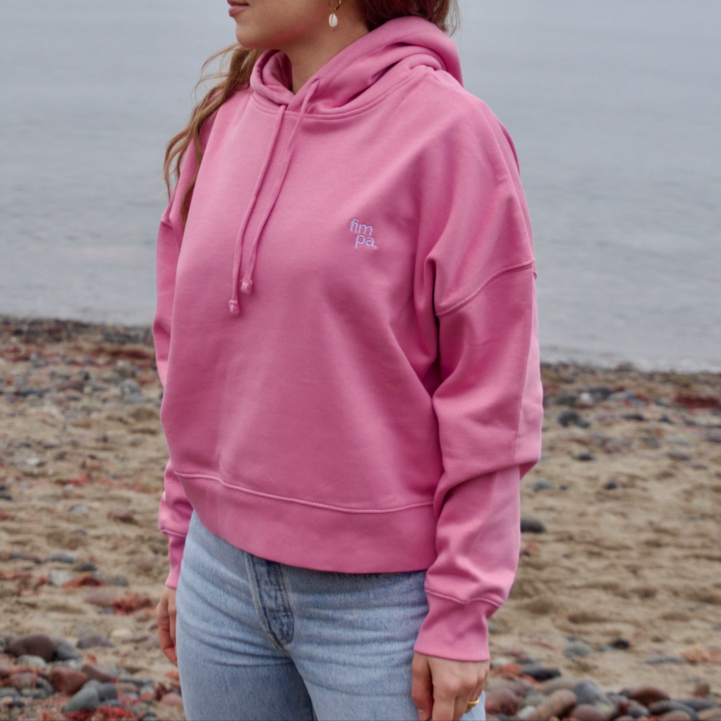 Fairer Hoodie in Rosa bzw. Pink, getragen von einer Frau am Strand, nachhaltige Mode von fimpa.de.
