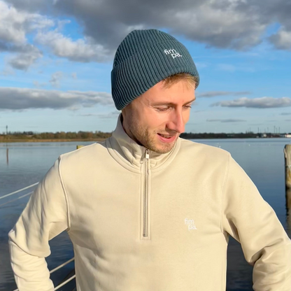 Person trägt einen petrolfarbenen fimpa-Beanie und einen beigen Pullover mit Reißverschluss vor einer malerischen Kulisse am Wasser – moderner und nachhaltiger Look.