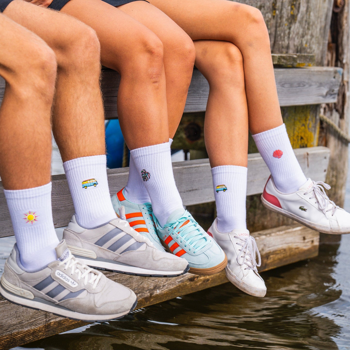 3 Personen präsentieren ihre weißen Socken mit verschiedenen dezenten und grellen Stick-Motiven. Sie sitzen auf einem Steg und lassen die Füße über dem Wasser baumeln.