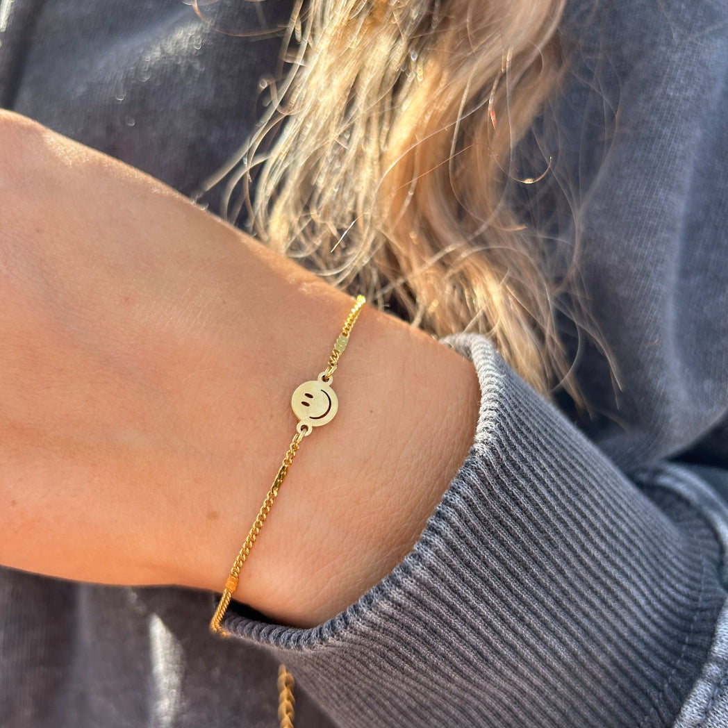 Trendiges 18 Karat goldenes Smiley Armband als Accessoire mit einem schwarzen Used-Look Pullover kombiniert.