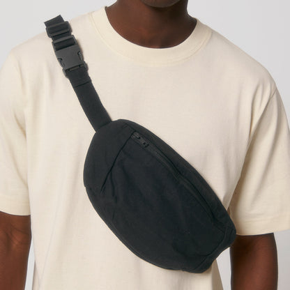 Mann in einem cremefarbenen T-Shirt trägt eine schwarze Bauchtasche aus nachhaltigem Material quer über die Schulter. Die Tasche verfügt über einen dezenten Reißverschluss und einen verstellbaren Trageriemen mit Clipverschluss.
